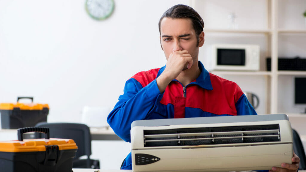 A professional AC repairman visibly reacting to a foul odor while inspecting an air conditioning unit, illustrating the article's subject of unusual smells emanating from AC units.