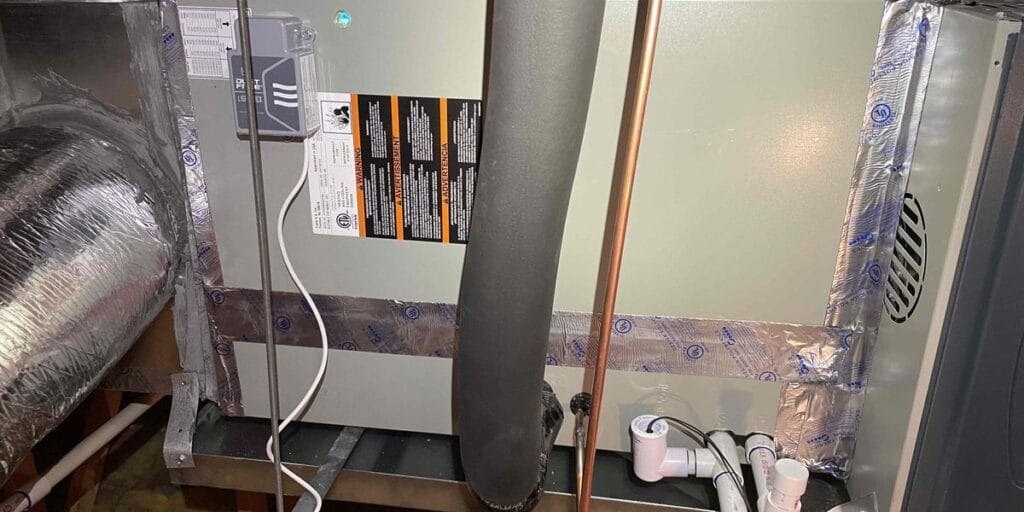 An indoor air handler unit sits in an attic with a bare copper refrigerant line and white PVC condensate pipes running alongside it.