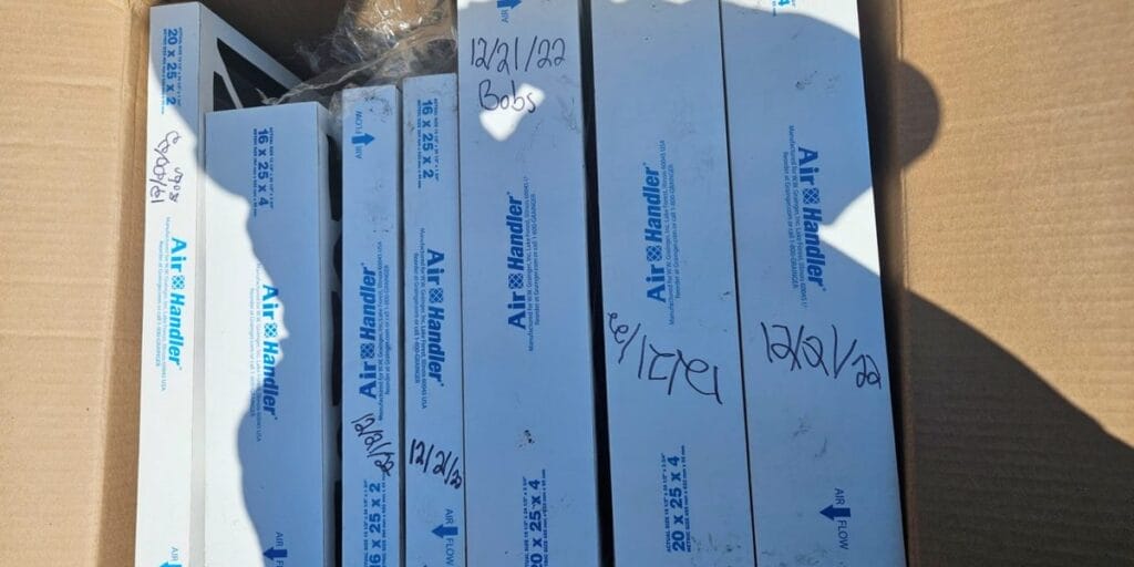 A cardboard box holds several new replacement air filters, each hand dated in marker, with a shadow cast across them.