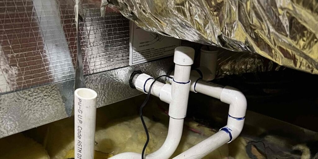 White PVC pipes with several elbow joints run beneath an insulated air handler in an attic