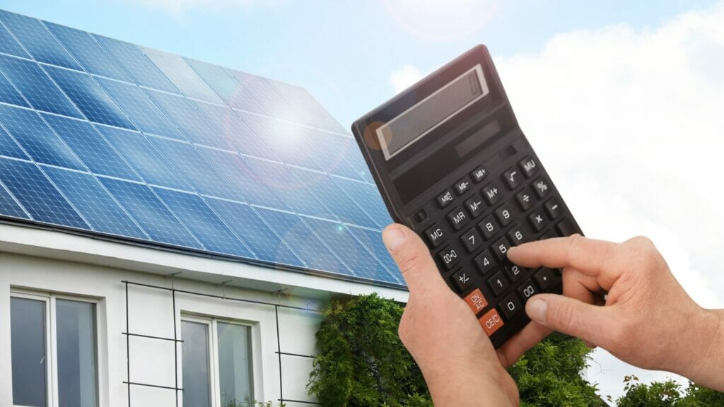 Man using a calculator to calculate savings, with a house featuring a solar panel-covered roof in the background