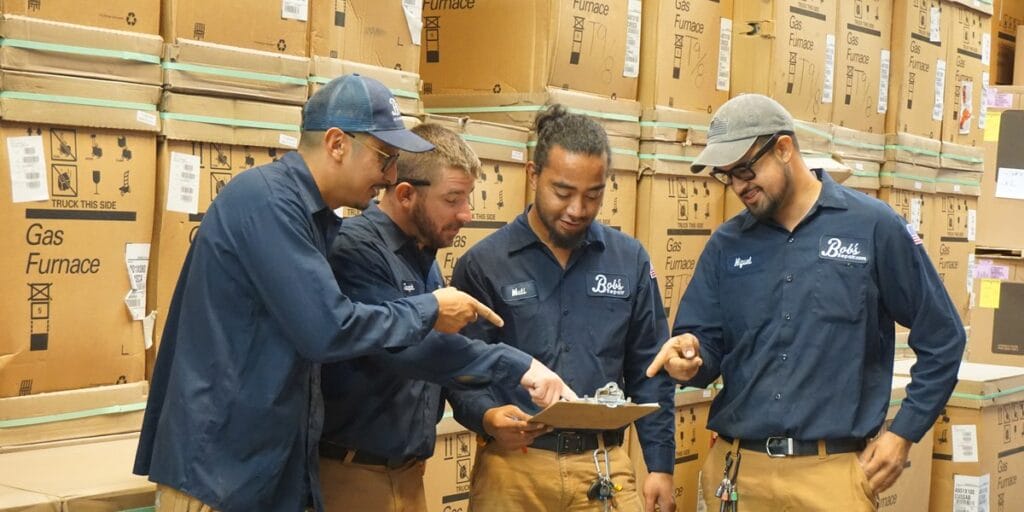 Four Bob's Repair technicians in matching uniforms examine a small part on a clipboard inside a warehouse stacked with furnace boxes.