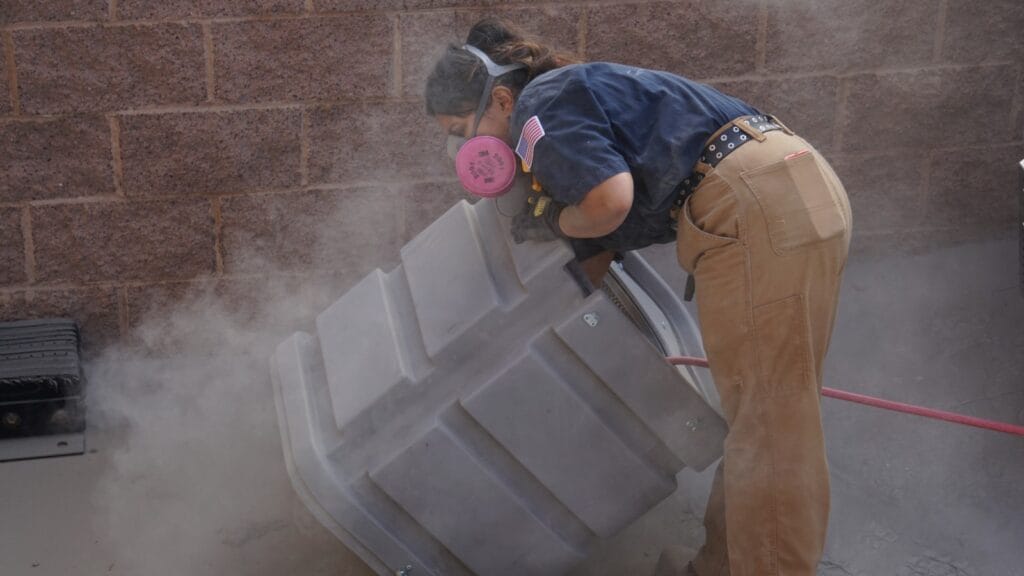 This image shows a worker from Bob's Repair engaged in the process of air duct cleaning. The worker is wearing a navy blue uniform shirt and light brown work pants and is equipped with a pink respirator mask, ensuring protection from dust and other particulates. The worker is bending over a large, grey air duct, which is emitting a cloud of dust or debris, suggesting active cleaning