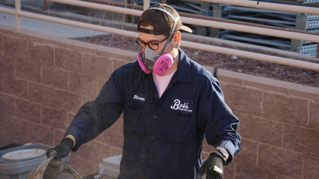 A Bob’s Repair employee is performing outdoor maintenance work. He is outfitted in a navy blue work uniform with the company logo visible on his shirt. Safety is prioritized with his personal protective equipment, which includes a gray mask with pink filters over the nose and mouth and protective gloves.