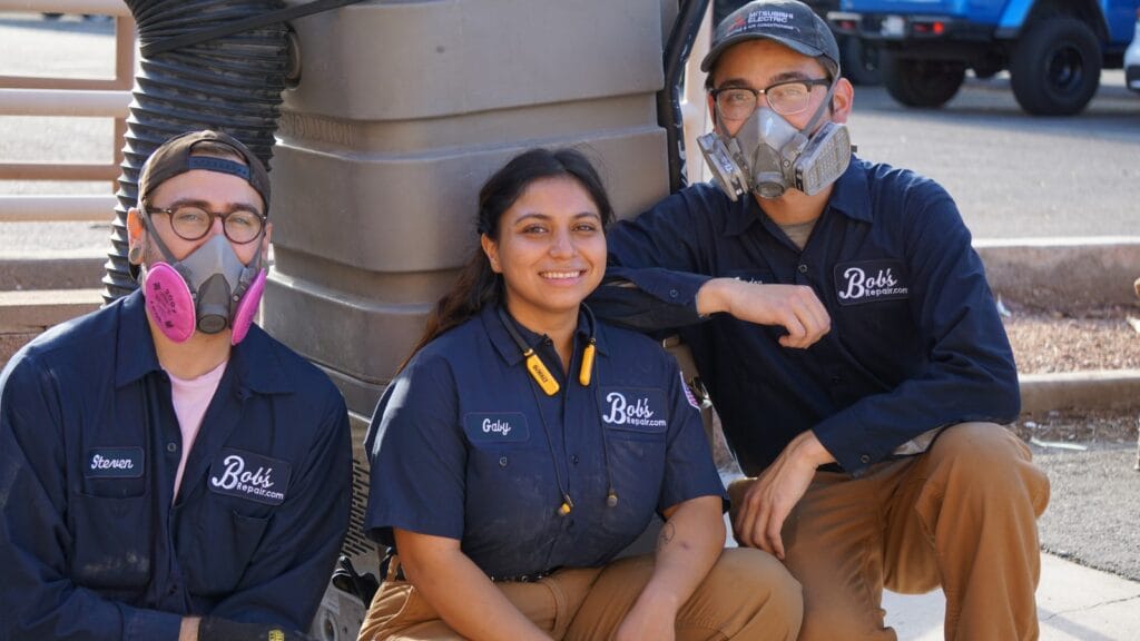 Three HVAC technicians wearing Bob's Repair uniforms are sitting together, smiling after completing an air duct cleaning job. The two individuals on the sides are wearing protective respirator masks, and all three have name tags. They are seated in front of a large, grey air duct system, which suggests the environment of their recent work.