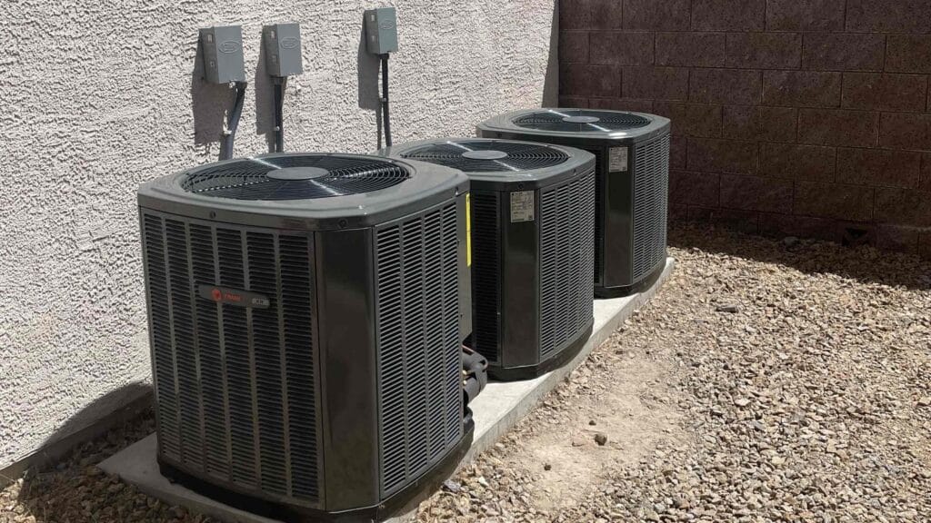 Three industrial air conditioning units on concrete pads against a textured stucco wall, with electrical boxes above on a sunny day.