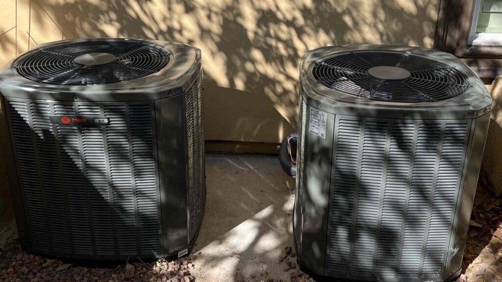 Two outdoor air conditioning units with horizontal vent slats and fan grilles on top, positioned side by side against a house wall with leafy shadows cast upon it.
