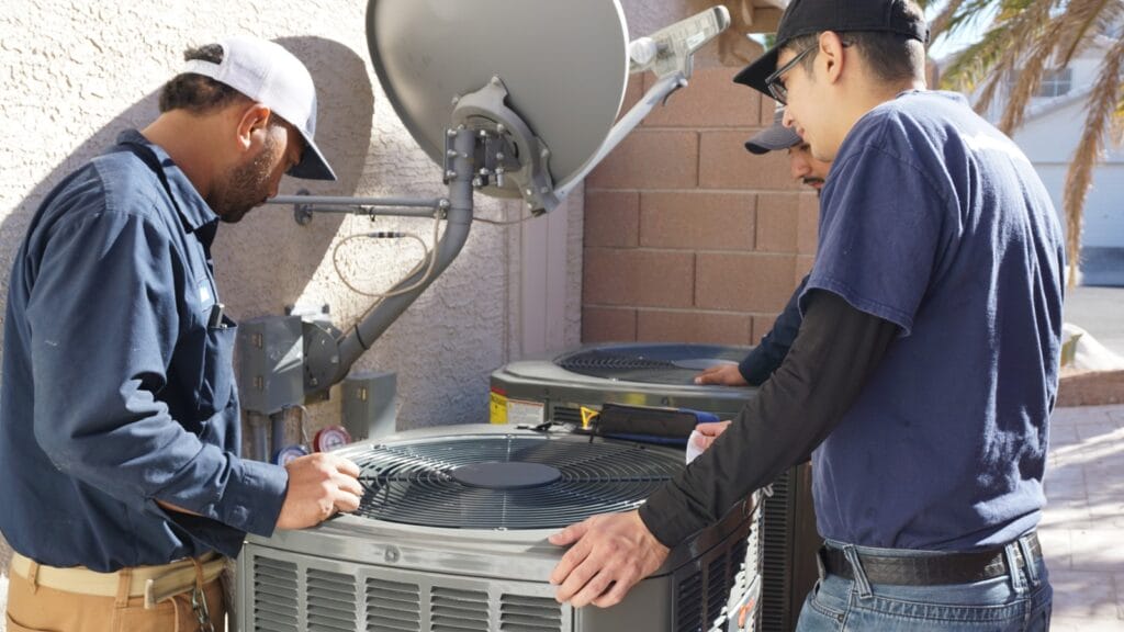 Two Bob’s Repair technicians servicing an outdoor air conditioning unit beside a residential building, illustrating an AC repair scenario.