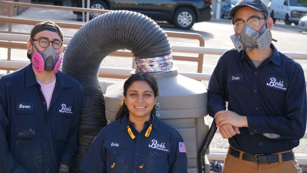 Three professional air duct cleaning technicians from Bob's Repair stand confidently in front of an HVAC system, with two wearing protective respiratory masks. They are dressed in company uniforms, indicating their expertise in maintaining indoor air quality through specialized cleaning services.