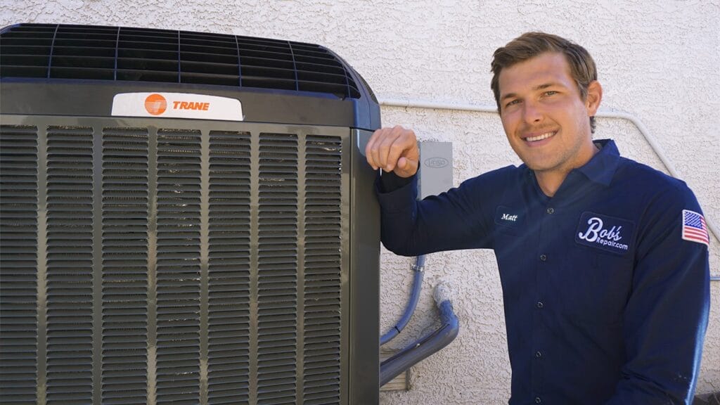 Smiling technician in a blue uniform labeled 'Bob's Repair.com', standing next to an outdoor HVAC unit, representing professional air conditioning service and maintenance.