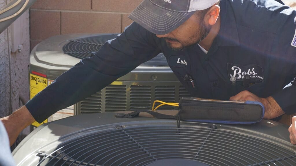 A technician named Luis, wearing a cap and a navy uniform labeled "Bob's Repair," is servicing an outdoor HVAC unit, with tools on hand for maintenance.
