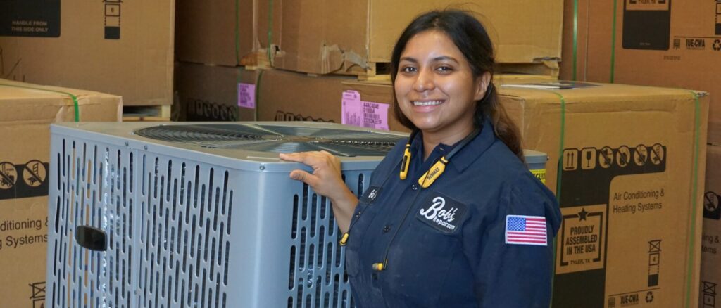 Technician standing next to an AC condenser unit in a warehouse.