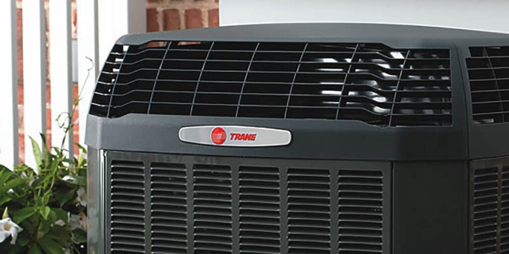 A close-up of a dark gray Trane air conditioning unit with a red and white logo badge on the front.