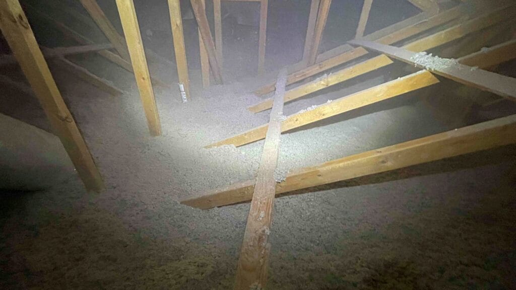 An attic covered with loose-fill insulation, showing wooden beams and a dimly lit environment. Insulation appears freshly blown into the space.