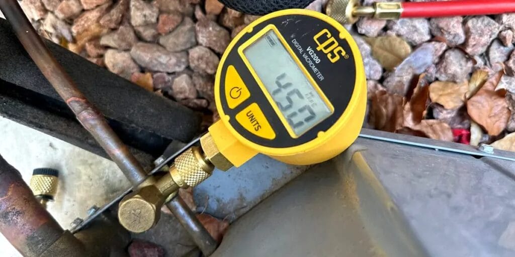 A yellow CPS digital vacuum gauge with a round display reading 45.0 rests on a copper refrigerant line, sitting on gravel landscaping near an outdoor condenser unit.