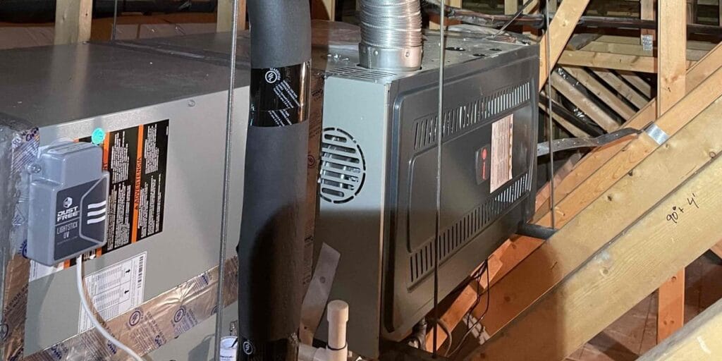 A gas furnace sits tucked into a tight attic space with a metal flue pipe and insulated line running up through the wood framing