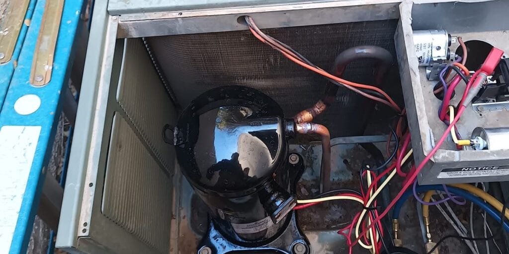The inside of an outdoor condenser cabinet is exposed showing the black compressor, copper refrigerant lines and electrical wiring