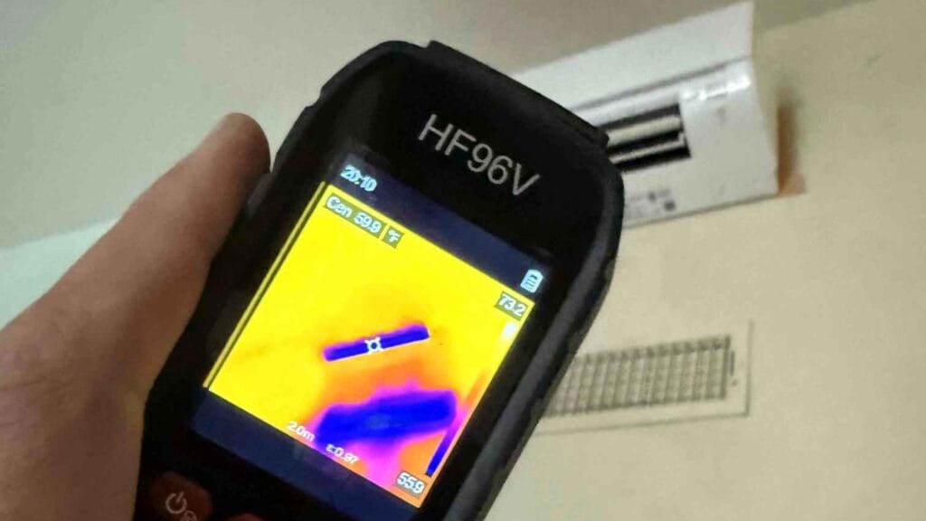 HVAC technician using a thermal imaging camera to check an indoor AC unit and vent for temperature anomalies or air leaks during maintenance.
