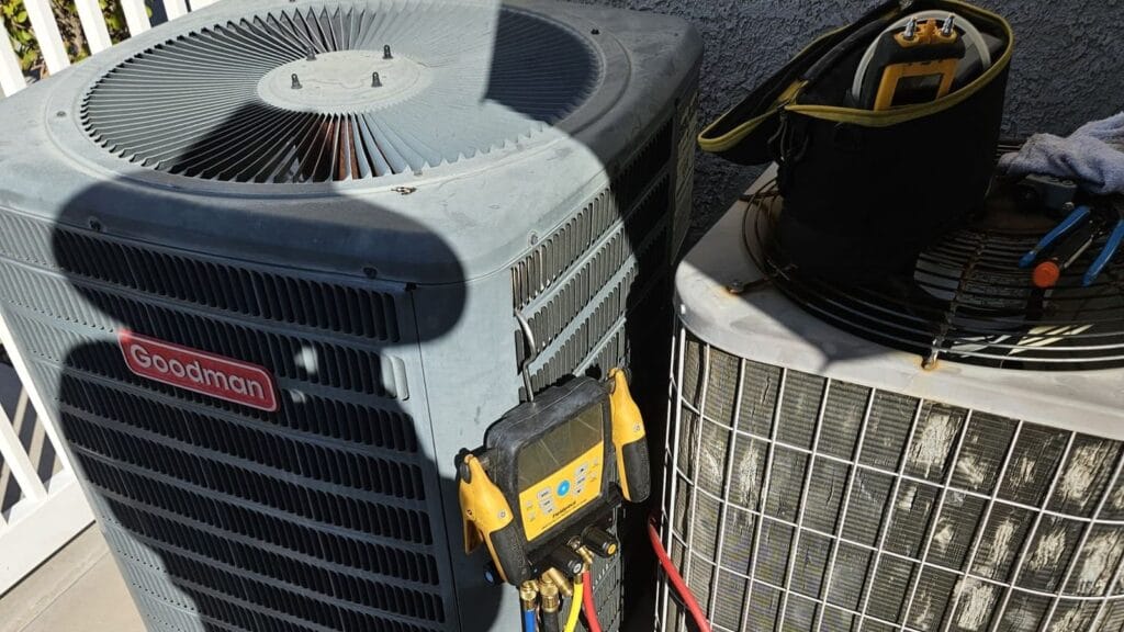 A Goodman outdoor condenser unit connected to a yellow refrigerant gauge sits beside a second condenser unit whose coils are visibly caked with dust, with a tool bag and pliers resting on top.