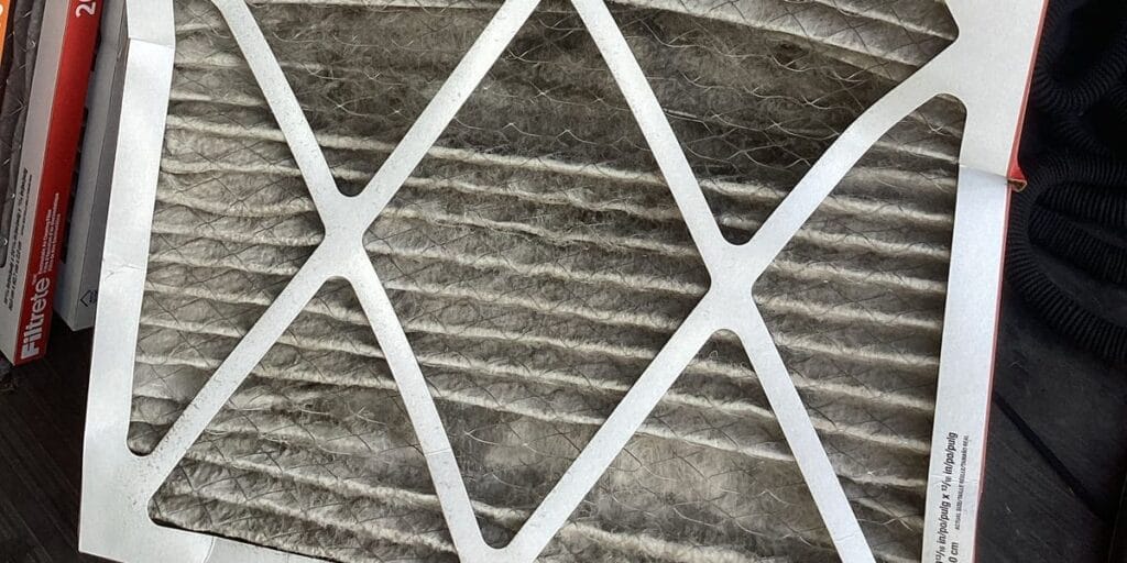A dirty gray pleated air filter leans against a stack of new Filtrete filter boxes on the floor.