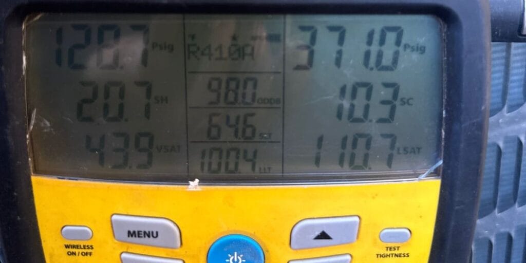 A yellow Fieldpiece digital refrigerant gauge displays several pressure and temperature readings on its screen, mounted next to an outdoor unit.