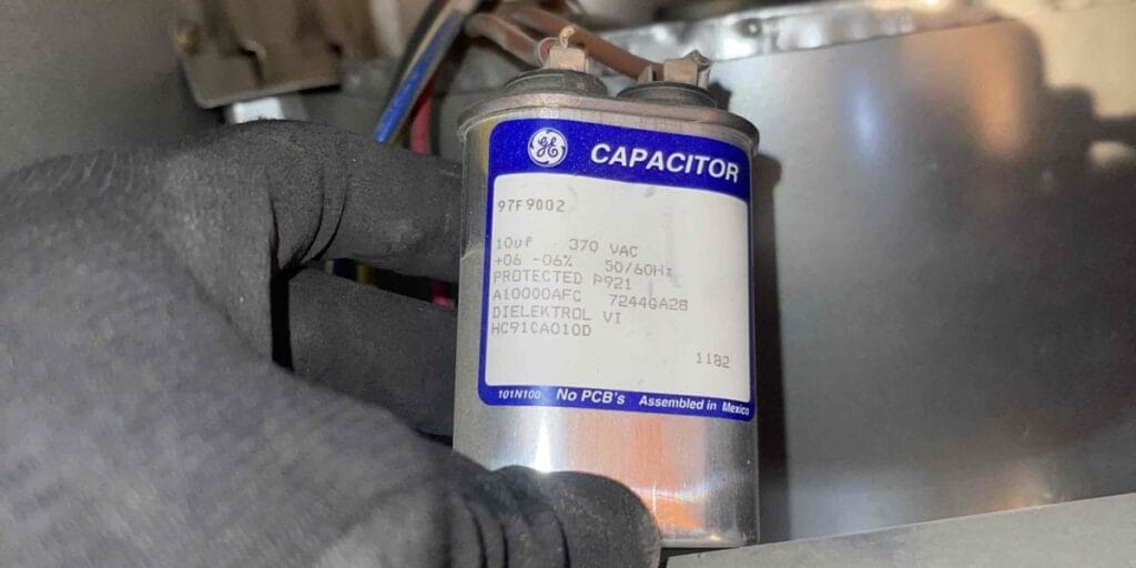 A gloved hand holds a cylindrical GE capacitor with a blue label, still connected by wires inside an air conditioning unit's housing.