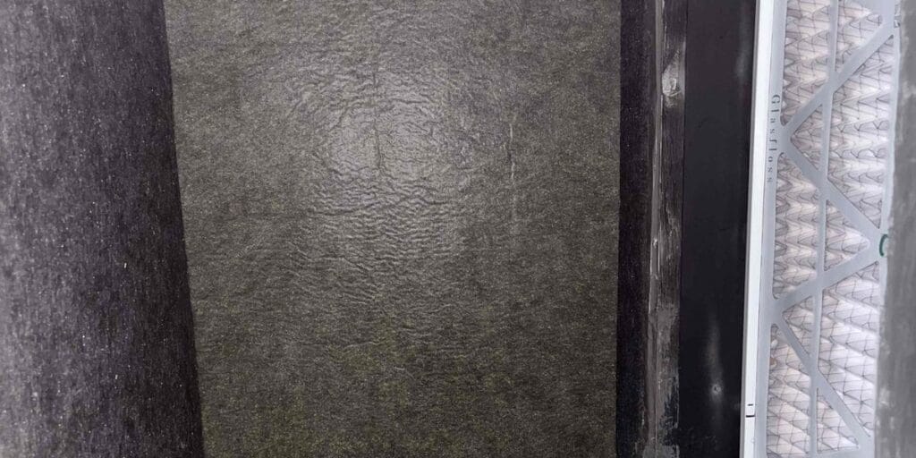 The inside of an air handler cabinet with dark grimy insulation lining next to a clean replacement filter.