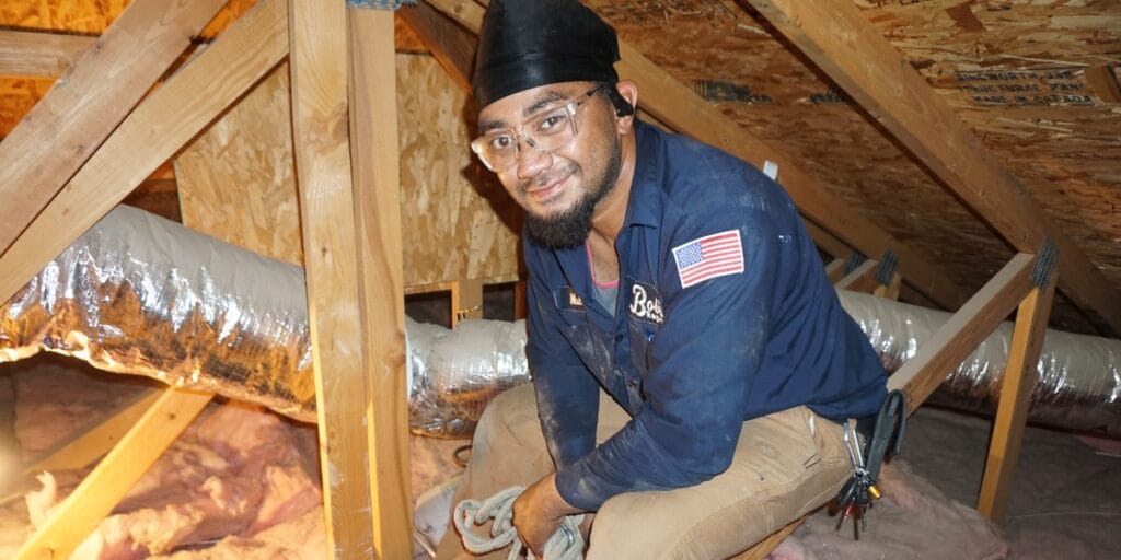 A Bob's Repair technician kneels in an attic surrounded by ductwork and insulation, holding a coiled rope.