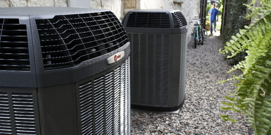 Two outdoor AC condenser units line a narrow gravel side yard with a person walking past in the background.