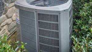 An outdoor air conditioning condenser unit surrounded by shrubs against a stone wall.