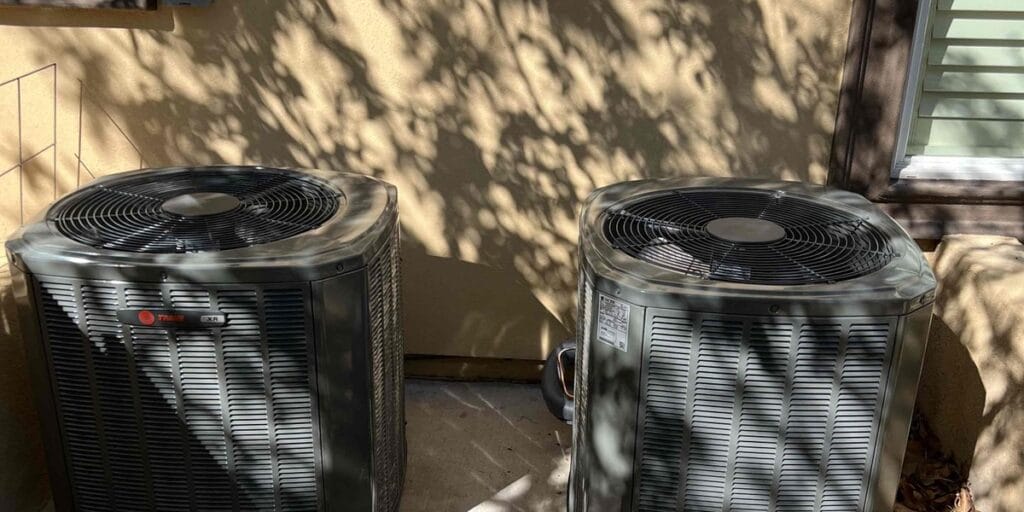 Two outdoor air conditioning condensers sit close together in a shaded side yard.