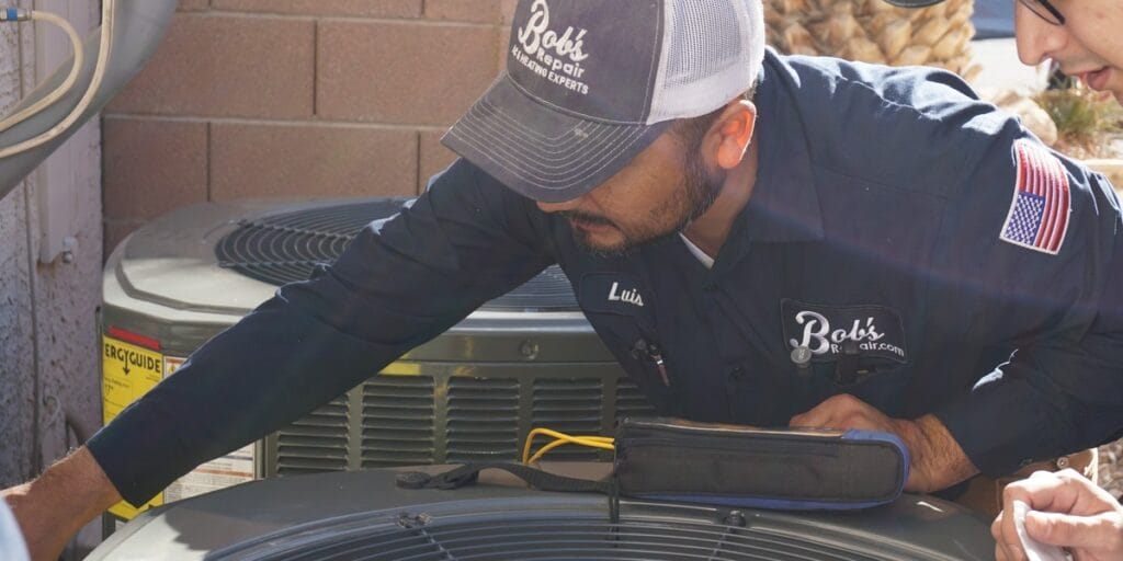 A Bob's Repair technician wearing a company cap and uniform leans over an outdoor condenser unit, checking its components with a tool bag open beside him.