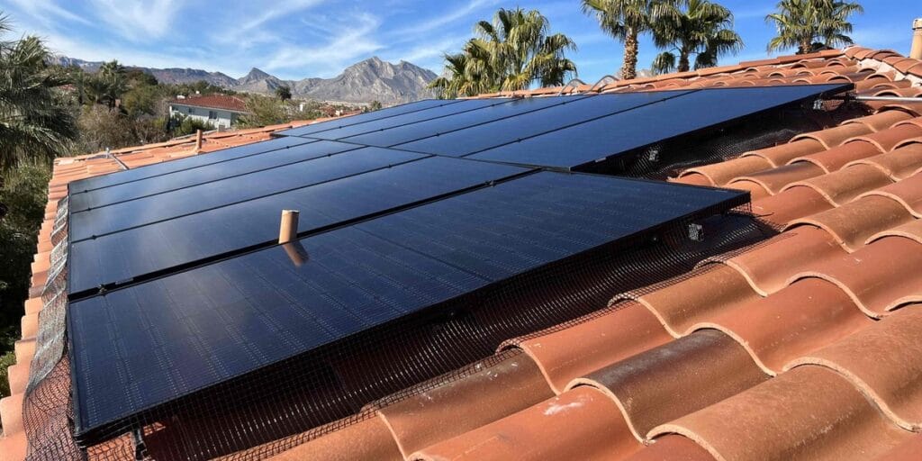 A large solar panel array covers a red tile roof, framed by desert mountains and palm trees under a clear blue sky.