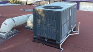 A rooftop package AC unit sits on a flat red membrane roof next to old insulated ductwork, with a neighboring garage visible in the background.