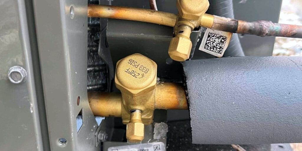 A close-up of brass shutoff valves and copper refrigerant lines on the side of an outdoor AC unit, with insulated tubing running into the cabinet.