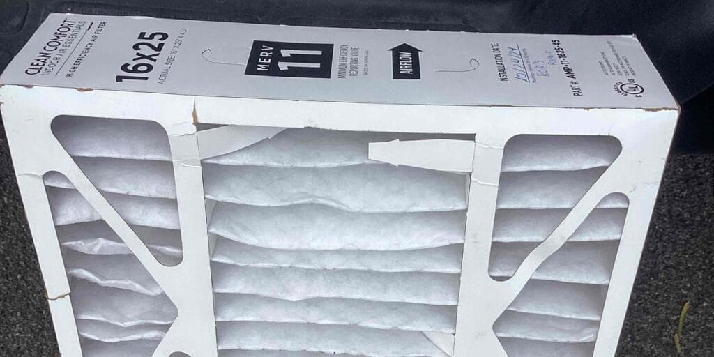 A brand new pleated air filter still in its box, marked 16x25 and MERV 11, sitting on a driveway.