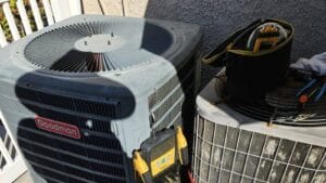 Two outdoor air conditioning units side by side with diagnostic gauges and a tool bag set up for a repair