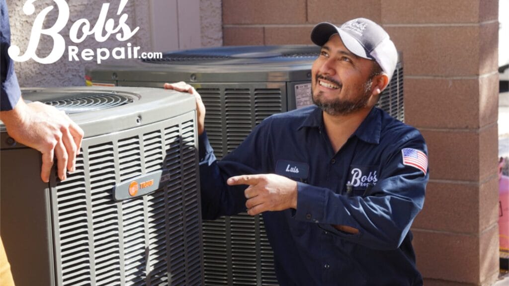 A smiling Bob's Repair technician in a dark blue uniform points cheerfully at an outdoor heating and cooling unit while talking to someone off camera.