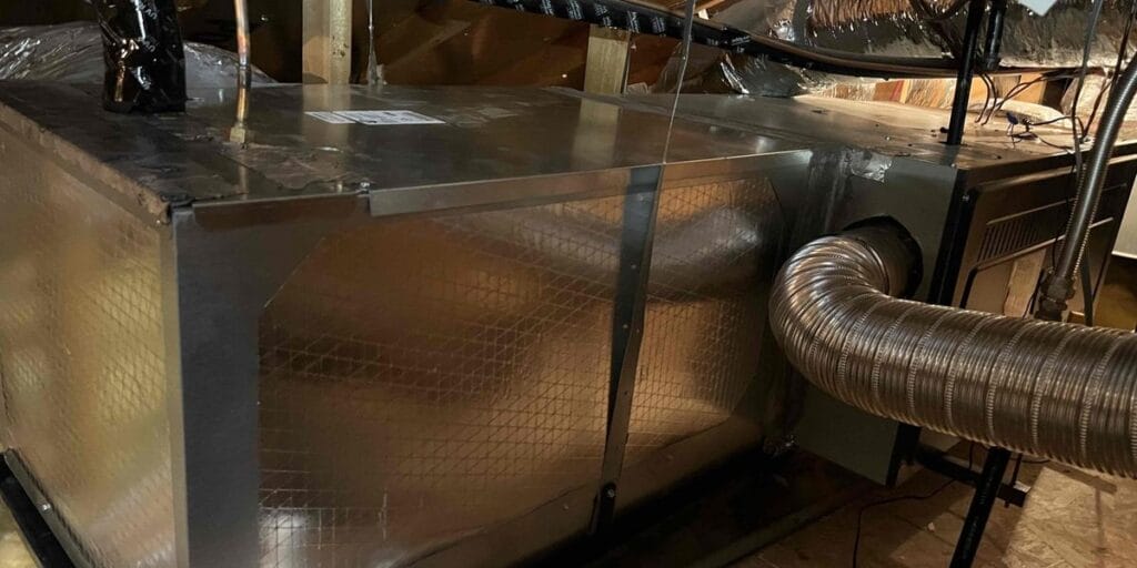 A large rectangular metal heating unit tucked away in a wooden attic with a flexible ribbed metal exhaust pipe curving out from the side.