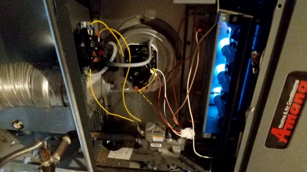 An open furnace cabinet showing complex wiring and a motor on the left with bright blue flames burning safely inside the right side of the unit.