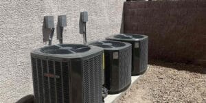 Three outdoor AC units lined up against a stucco wall, each with its own electrical disconnect box mounted above it in a gravel side yard.