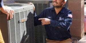 Bob's Repair technician Luis kneeling by two outdoor Trane units, pointing at one and smiling while another tech's hand rests on top