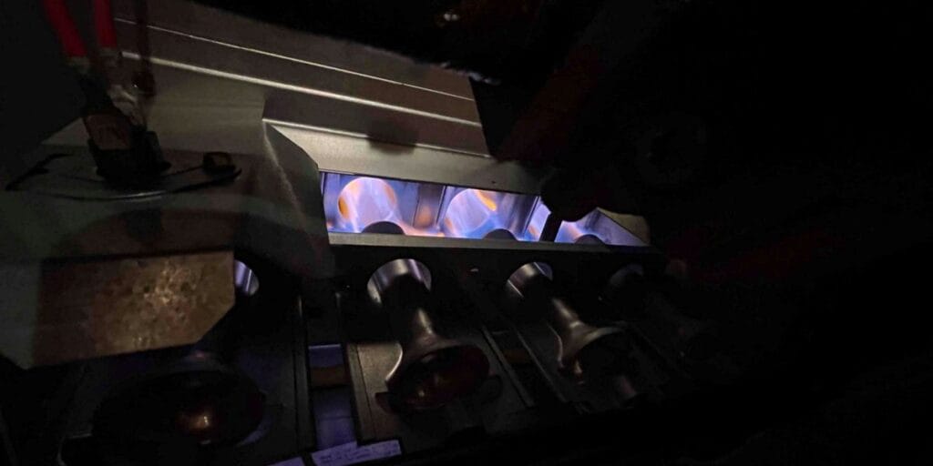Close-up inside an open furnace cabinet showing several gas burners lit with steady blue flames in a dark compartment