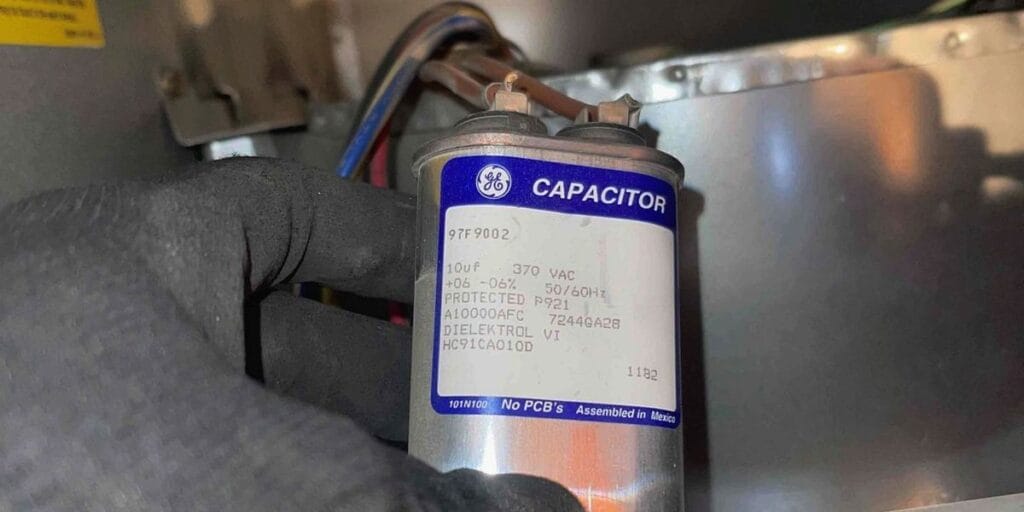 A gloved hand holding a GE capacitor for an HVAC system during an AC maintenance check in a Las Vegas home.