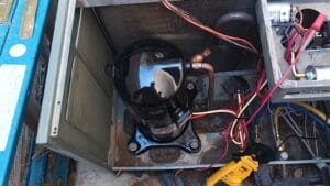 Open outdoor heat pump unit showing the black compressor, wiring, and a yellow clamp meter resting by the gauge hoses